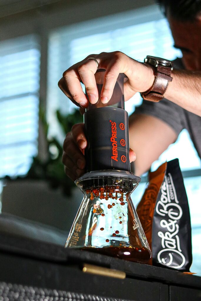Mãos pressionando um AeroPress sobre um recipiente de vidro, com gotas de café caindo. Um pacote de café está ao fundo, destacando o preparo manual de café.