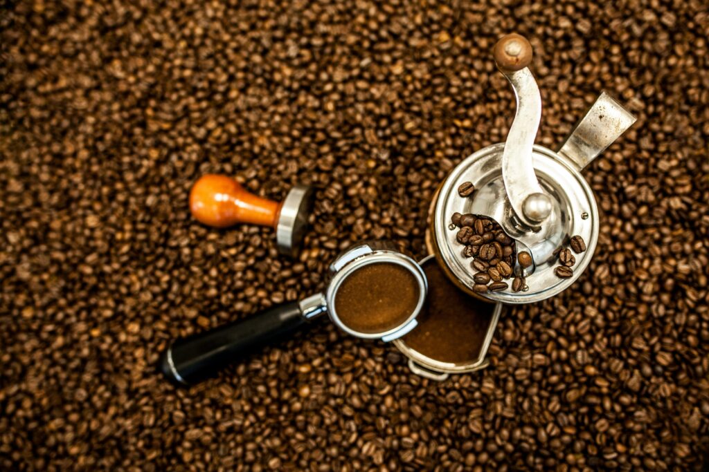 Imagem mostrando a preparação de café com um moedor manual, grãos de café, e um portafiltro sobre uma superfície coberta de grãos de café. O moedor manual está parcialmente cheio de grãos de café e o portafiltro contém café moído, destacando os elementos essenciais para o preparo do espresso.