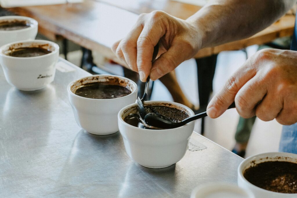 Mãos segurando colheres sobre xícaras brancas durante uma sessão de degustação de café, com várias xícaras alinhadas em uma mesa de metal.