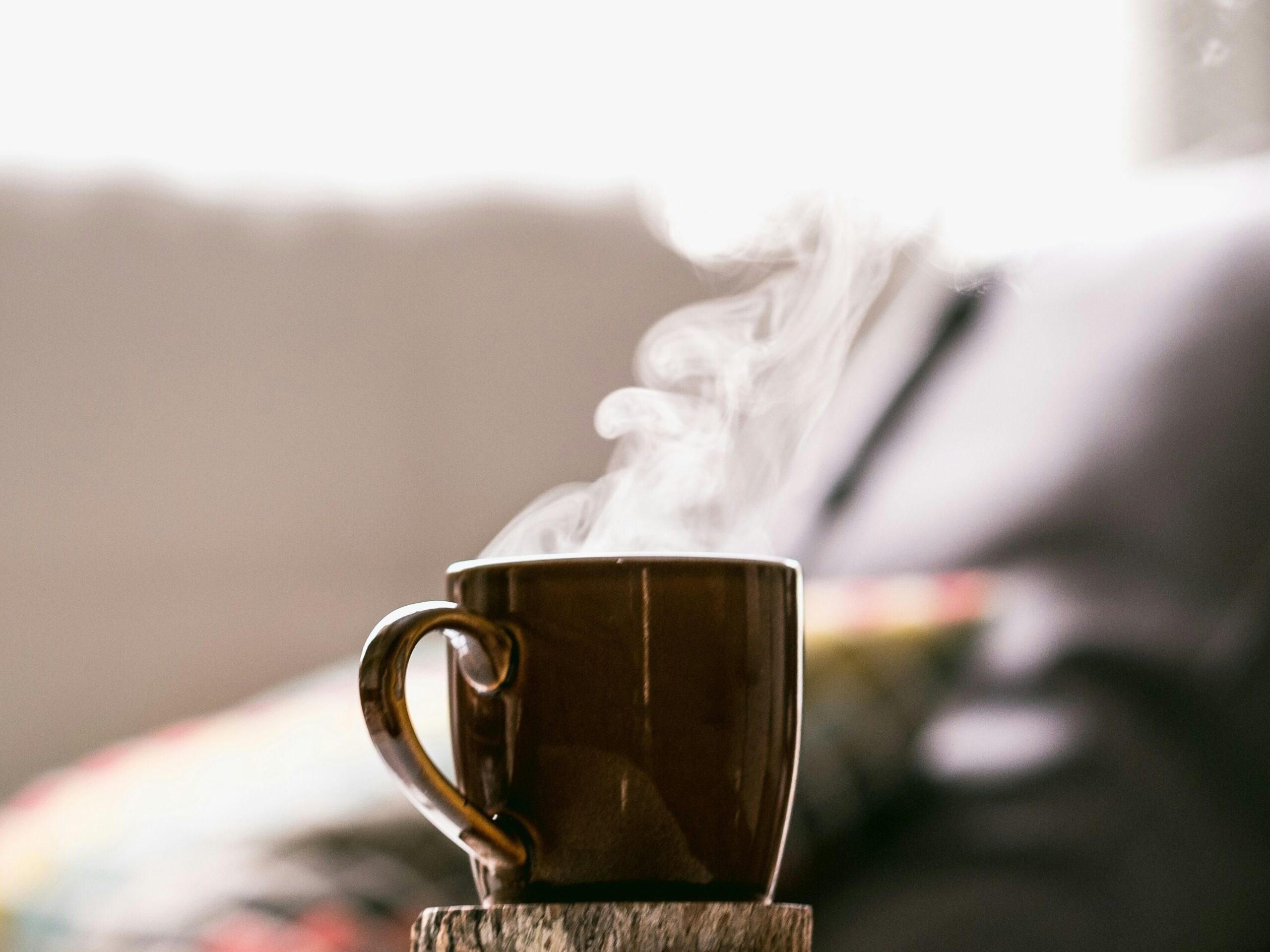 Uma caneca de café marrom com vapor subindo, destacando-se em um ambiente acolhedor e desfocado ao fundo.